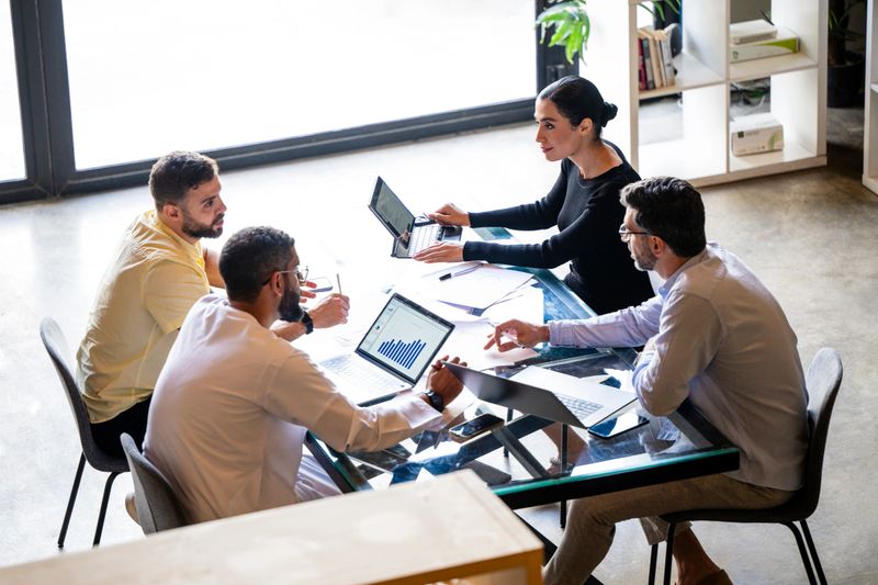 Three businessmen and one businesswoman sitting at table with computers, discussing in business meeting, teamwork, busy, working