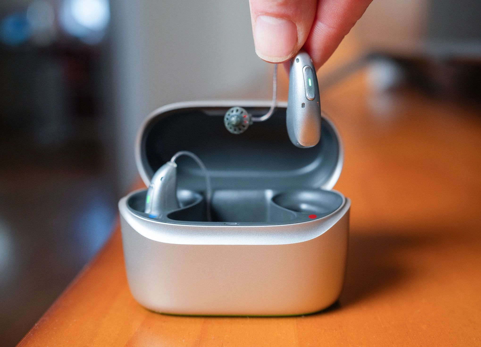 A hearing aid being held above its charging case on a wooden surface.