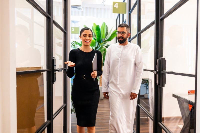 Businesswoman in her 20s holding laptop and gesturing,  businessman in his 30s wearing thawb, on way to meeting room