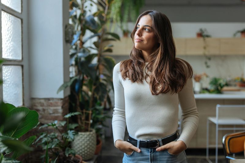 Serene young woman standing by the window, enjoying the natural light in her plant-filled home, contemplating the view