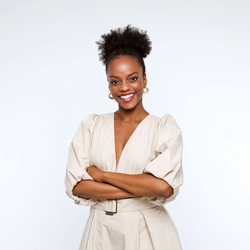 Confident and elegant afro american businesswoman wearing beige dress standing with arms crossed and smiling at camera. Studio shot, white background.