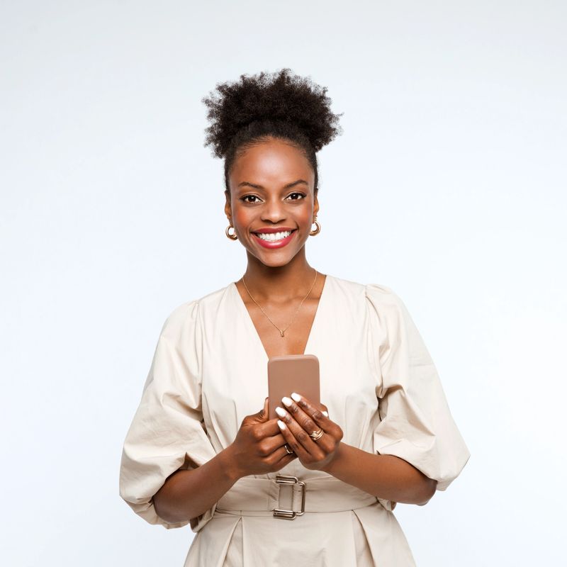 Confident and elegant afro american businesswoman wearing beige dress holding mobile phone in hands and smiling at camera. Studio shot, white background.