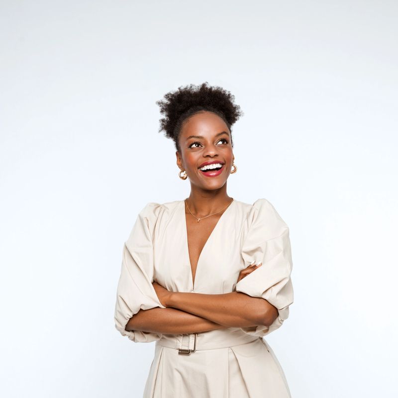 Excited afro american businesswoman wearing beige dress standing with arms crossed, looking away and smiling. Studio shot, white background.