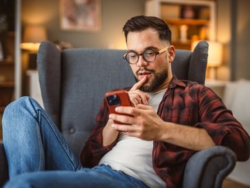 Man in glasses and plaid shirt sitting on armchair, looking thoughtfully at his phone.