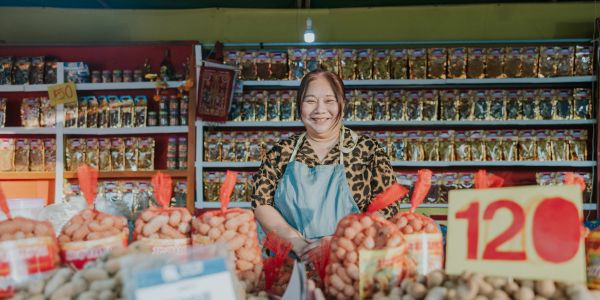Smiling vendor at a market stall with snacks and price signs.