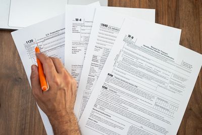 A person filling out U.S. tax forms with an orange pen on a wooden table.