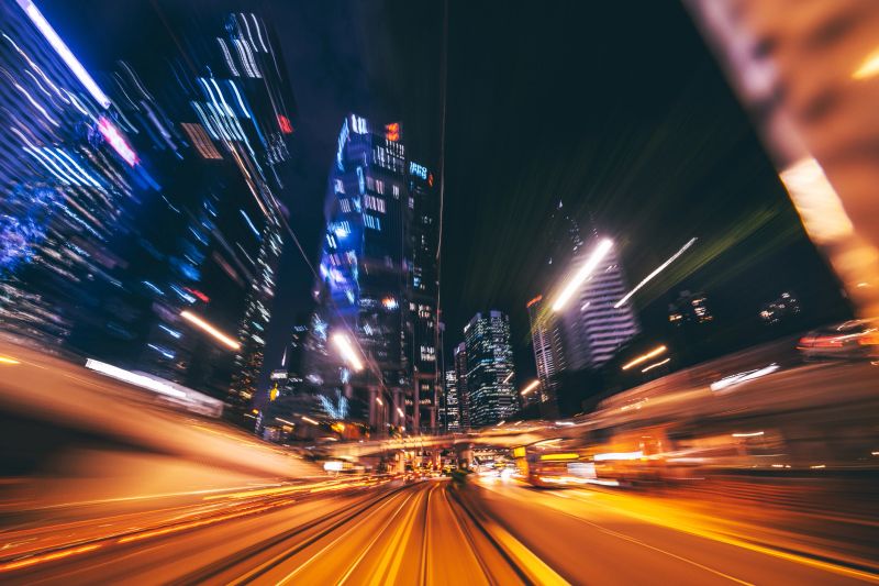 A vibrant depiction of the bustling city streets of Hong Kong at night, showcasing streaking lights and towering illuminated skyscrapers, encapsulating the city's energy and continuous movement.