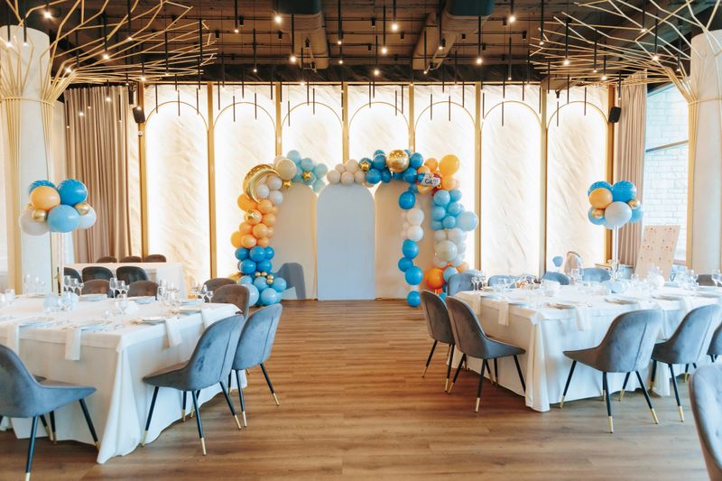 A beautifully decorated event venue featuring a balloon arch in blue, orange, and gold hues, elegant table settings with white tablecloths, and stylish modern chairs.