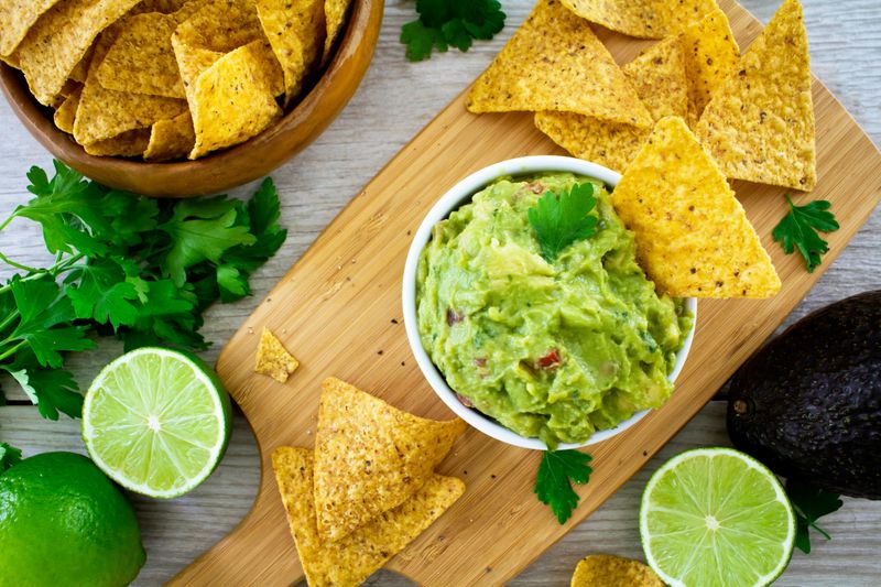 Fresh guacamole served in a bowl on a wooden chopping board, on a white wooden table. 
Shot from above with tortilla chips, fresh limes, avocado and sprinkled with parsley.
