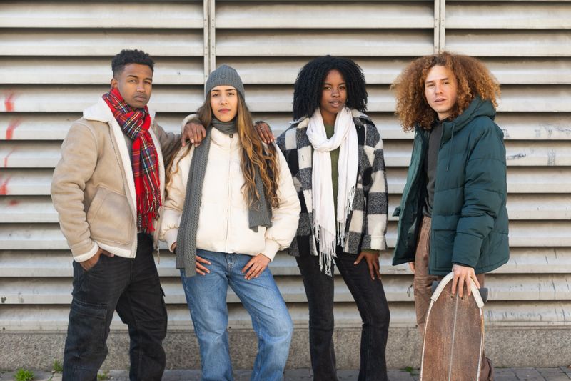 Four fashionable young friends are posing together on a city street, showcasing their trendy winter outfits and diverse styles
