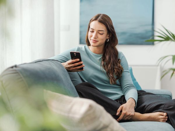 A woman relaxes on a couch, looking at her phone with a slight smile.