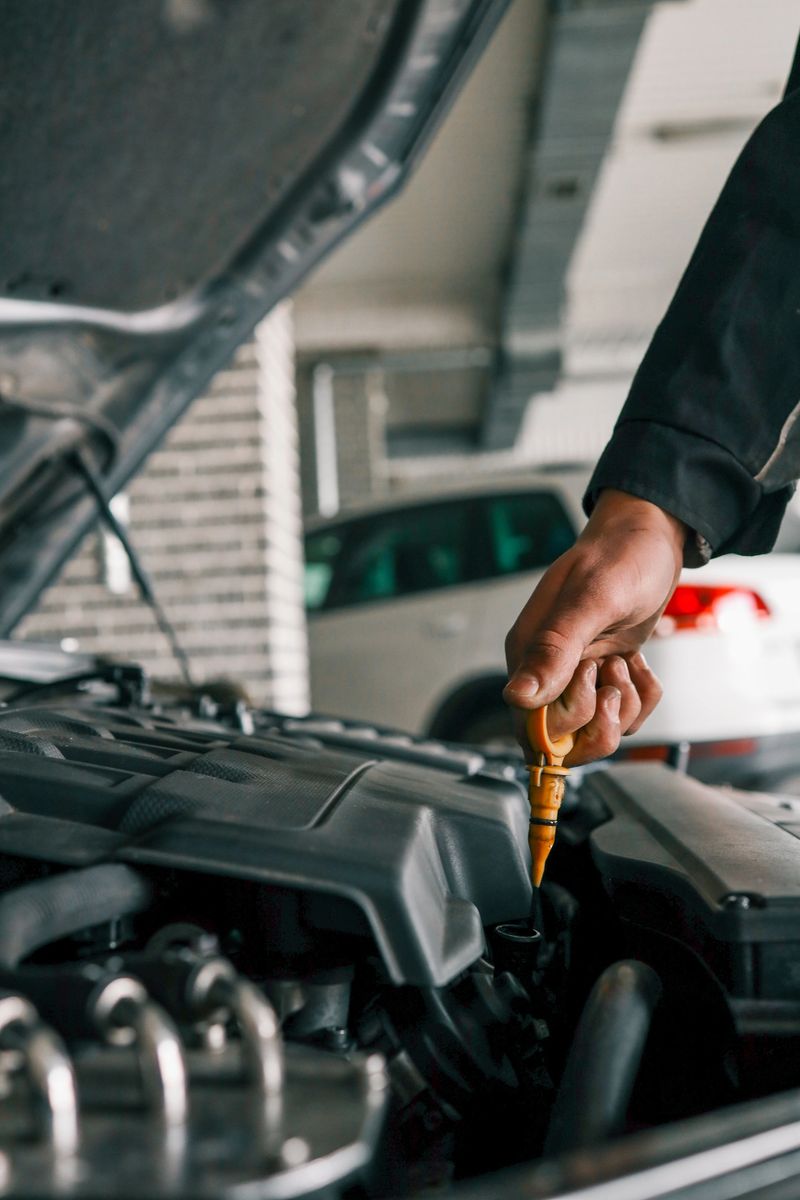 Close-up of a mechanic's hand holding an oil dipstick, checking engine oil levels in a vehicle's open hood. Represents routine car maintenance and servicing.