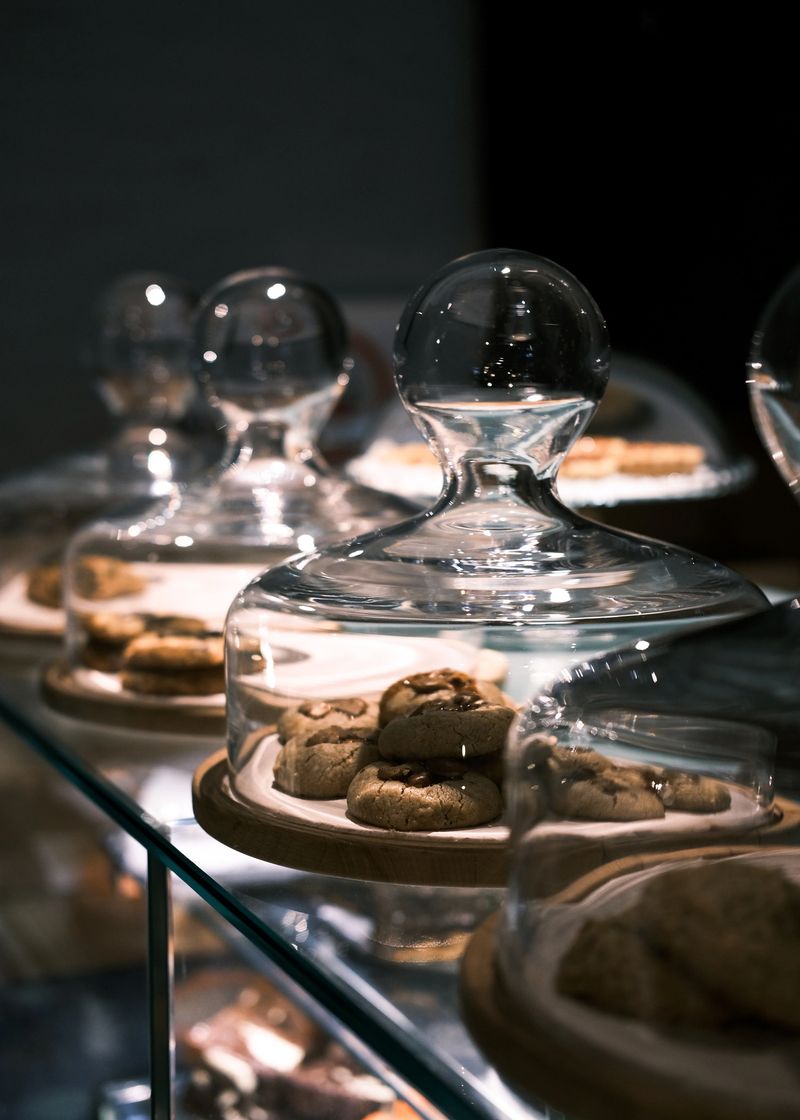 This image showcases an array of freshly baked cookies elegantly displayed under glass domes on wooden trays. The warm lighting highlights the golden hues of the cookies and the reflective surfaces of the glass covers, creating a luxurious and inviting atmosphere. Perfect for use in bakery branding, café menus, or promotional materials for baked goods, this composition emphasizes the craftsmanship and care in artisanal baking.