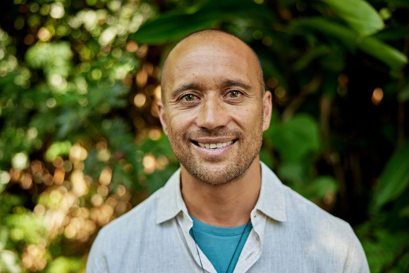 Close view of 46 year old individual with shaved head and beard stubble, wearing casual clothing and smiling at camera.