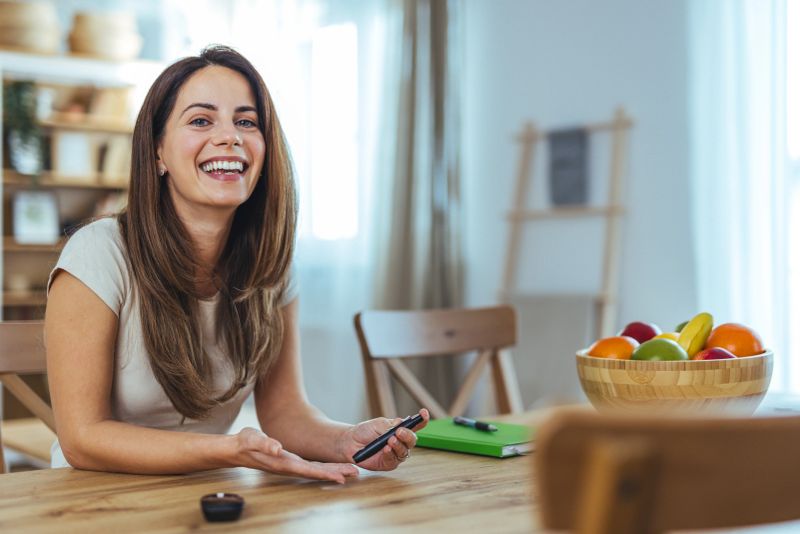 A cheerful woman is seated at a table, holding a smartphone, exuding joy and warmth. A bowl of colorful fruit is prominently displayed, symbolizing health and vitality.