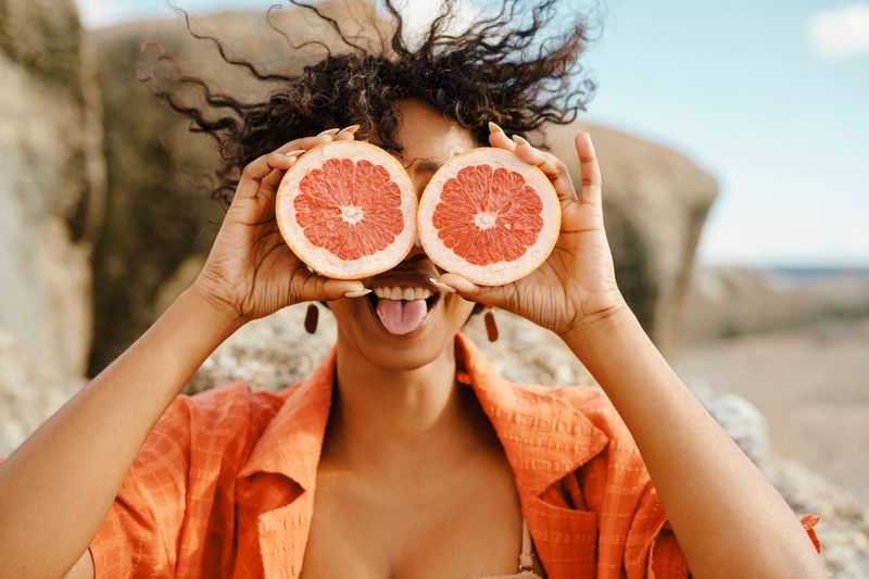A cheerful woman holding grapefruit halves in front of her eyes while outside in a sunny natural setting, exuding fun, happiness, and playful energy.