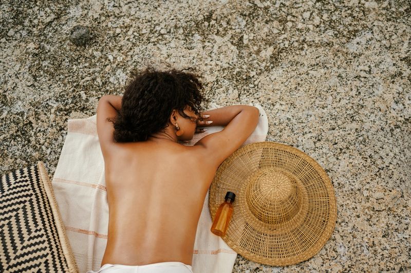 A relaxed outdoor scene of a woman resting on a towel on a stone surface, accompanied by a straw hat and summer beach essentials