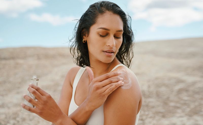 A young woman  applies sunscreen to her shoulder in a peaceful outdoor setting, highlighting sun protection and skincare
