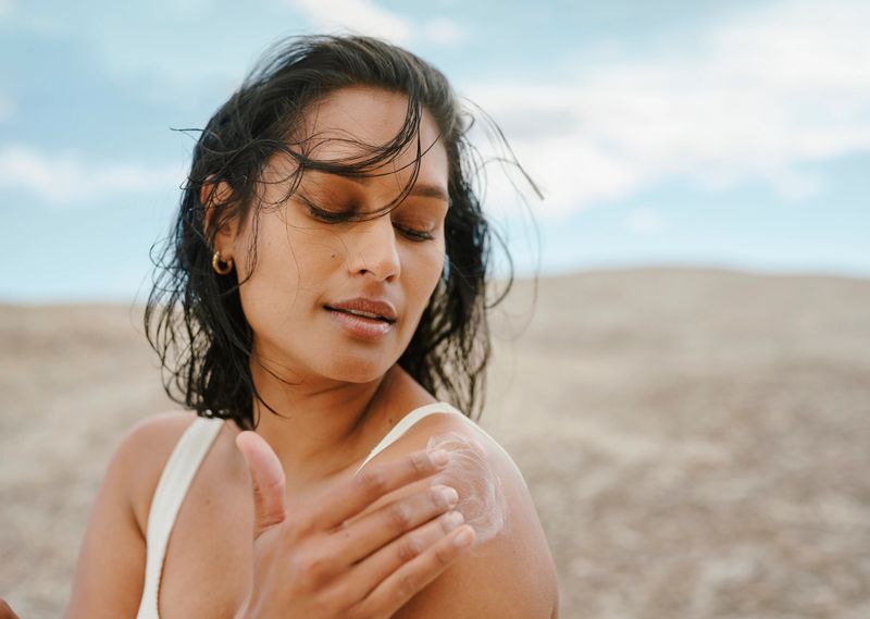 Close-up image of a woman applying sunscreen to her shoulder, set against a natural outdoor background and a clear blue sky.