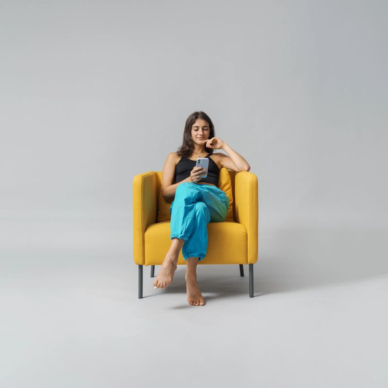 Happy stylish teenage girl sitting on an armchair and connecting with her smartphone, isolated on white background