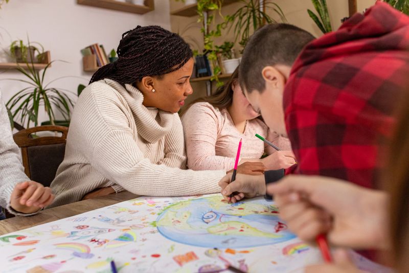 A group of diverse kids and their African American teacher come together around a table, joyfully working on an Earth Day poster.