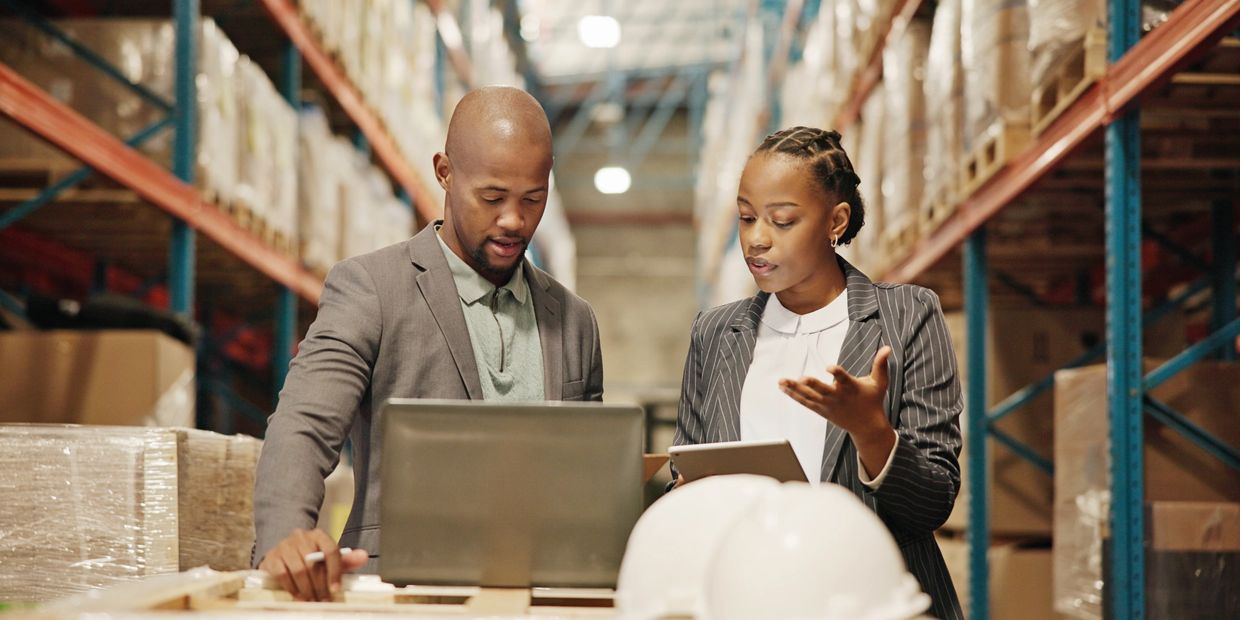 Two professionals discussing work in a warehouse with a laptop and tablet.