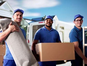 Three movers in blue uniforms carrying furniture and boxes outdoors.