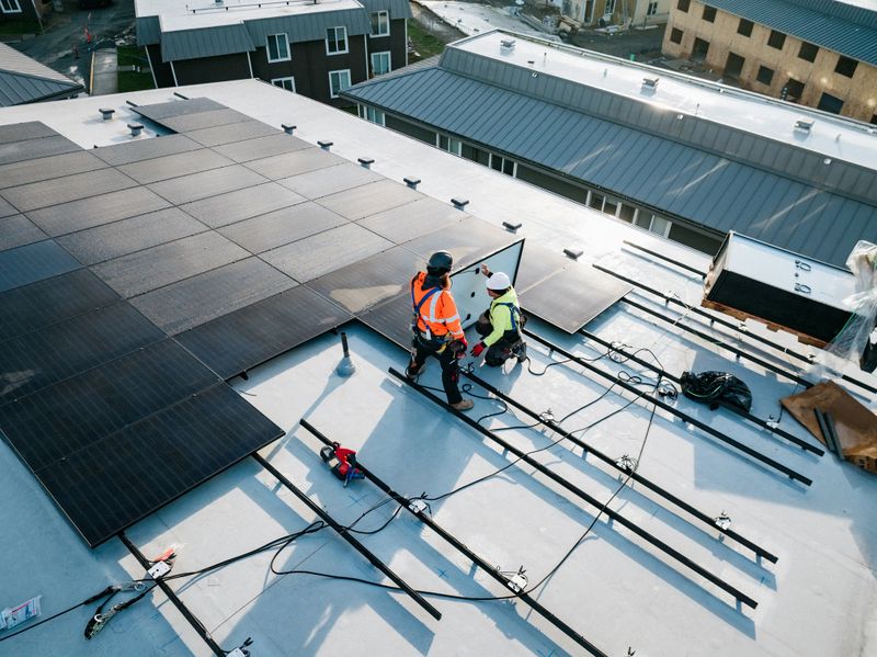 A group of PV Installers (Solar Photovoltaic Installers) work on the roof of an apartment complex, equipping one of a number of buildings to be energy efficient and environmentally friendly.  Shot from above with a drone.