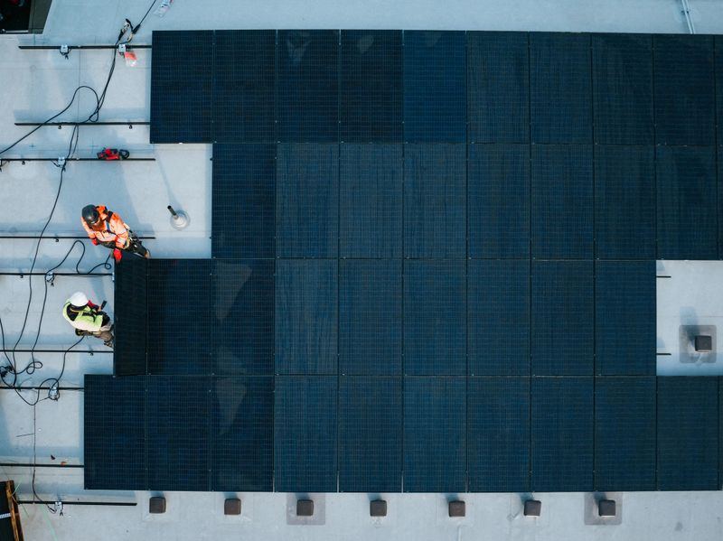 A group of PV Installers (Solar Photovoltaic Installers) work on the roof of an apartment complex, equipping one of a number of buildings to be energy efficient and environmentally friendly.  Shot from above with a drone.