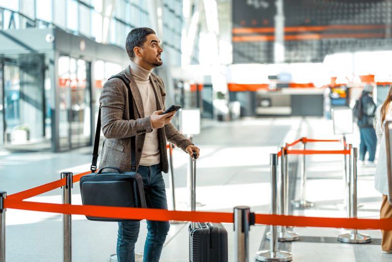 Businessman waiting in line at the airport terminal using his smartphone while holding his trolley suitcase and looking up to check the flight information