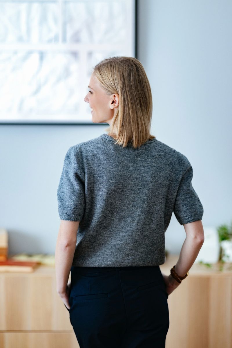 A young woman in professional attire stands in a modern interior, captured from behind. She wears a gray sweater and black pants, suggesting a focused and thoughtful professional demeanor in a creative workspace.