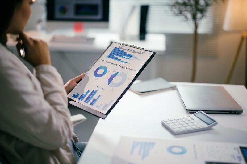 Businesswoman is analyzing financial charts and graphs on a clipboard, using a pen and sitting at her desk in a modern office, with a laptop and calculator nearby