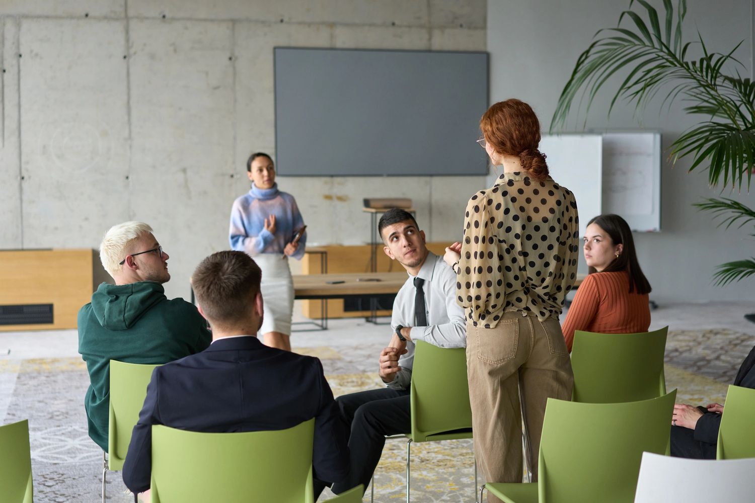 A woman standing and speaking to a seated group in a modern meeting room marketing her professional coaching services for communication training by communication skills coaching.