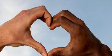 Two hands of different skin tones forming a heart shape against the sky.