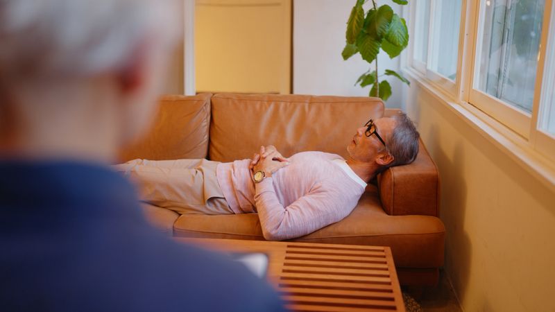 A senior man lies on a leather couch in a relaxed posture, engaging in a therapy session with a mental health professional who attentively takes notes. The serene environment, with warm lighting and greenery, promotes emotional well-being and mental health support.