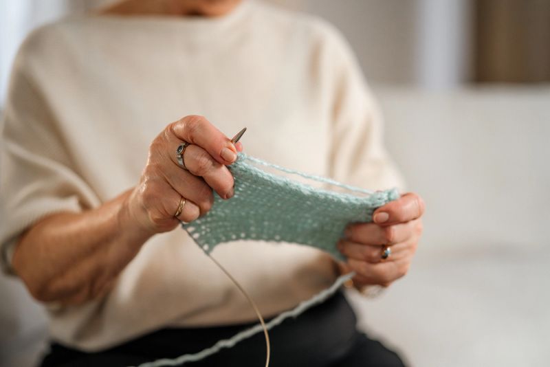A senior woman sits on a comfortable sofa, knitting with focus, in a cozy, well-lit living room environment. A laptop rests on a table, complementing the serene atmosphere.