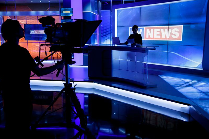 TV news studio ready for filming. Tv news anchor sitting in his place and two camera men silhouettes in front of him.