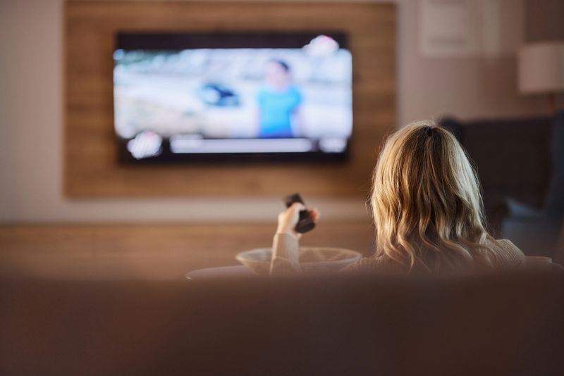 Rear view of carefree woman choosing what to watch on TV while relaxing on sofa in the living room. Copy space.