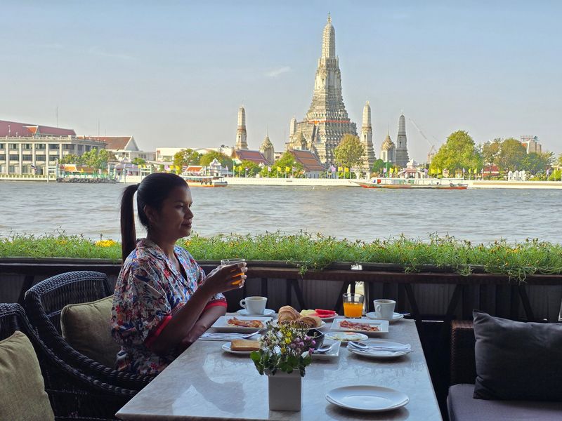 A woman savors her breakfast while overlooking the Chao Phraya River. Wat Arun stands majestically in the background on a clear morning in Bangkok, creating a peaceful atmosphere.