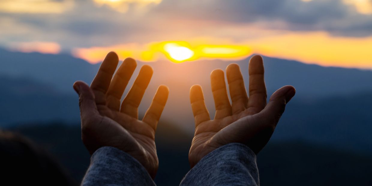 Hands reaching towards a glowing sunset over mountains.