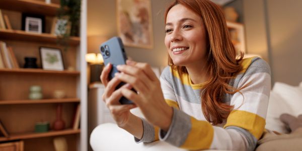 A smiling woman holding a smartphone while sitting comfortably indoors.