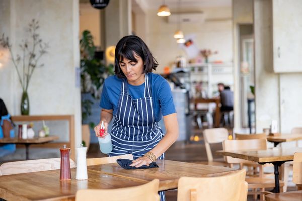 A woman cleaning a wooden table in a restaurant with a spray bottle and cloth.