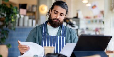 Man in apron reviewing documents at a laptop in a cozy workspace.