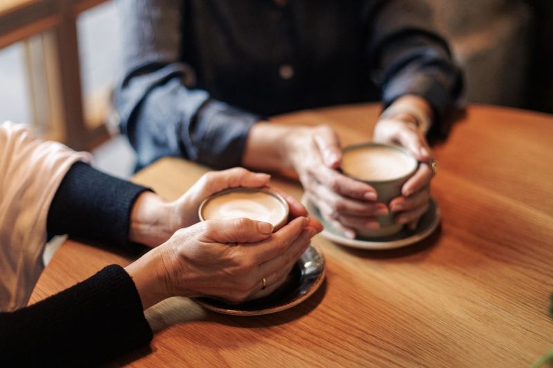 Two senior women enjoying warmth while holding cappuccino cups at a cafe table, sharing conversation and moments of comfort together