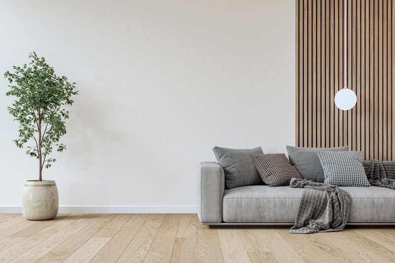 Modern interior / a living or waiting room with a gray sofa and decoration (a scattered blanket, pillows) on hardwood floor decorated with a pandant white lamp, and a large potted plant (a ficus benjamina plant) in front of a mainly white plaster partly hardwood paneled wall background with copy space. 3D rendered image.