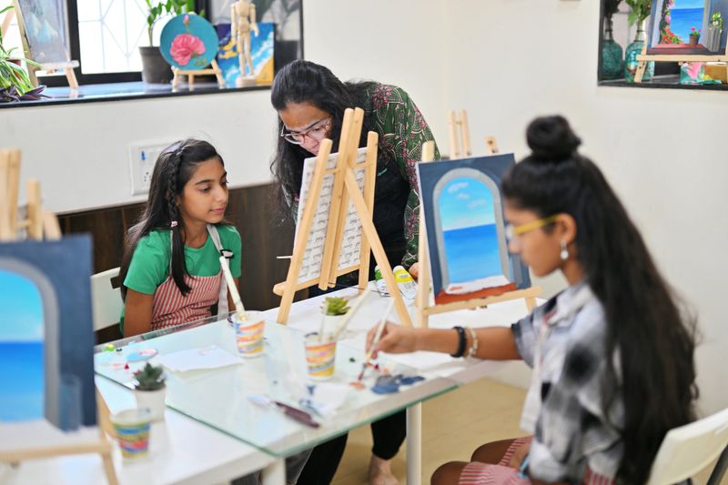 A skilled and passionate artisan is teaching a pre-teen girl the art of canvas painting in a bright, creative studio. The artisan demonstrates techniques with enthusiasm, showing the young girl how to apply brushstrokes and mix colors. The girl listens attentively, her face reflecting curiosity and excitement.