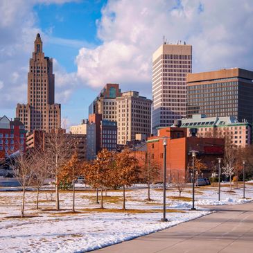 City skyline with snow-covered ground and autumn trees under a partly cloudy sky.