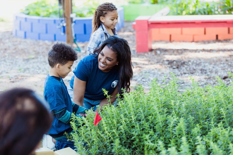 Smiling woman helps children plant herbs in an outdoor garden.  Community outreach and education.