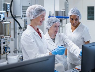 Three scientists in lab coats and hairnets working together in a lab.