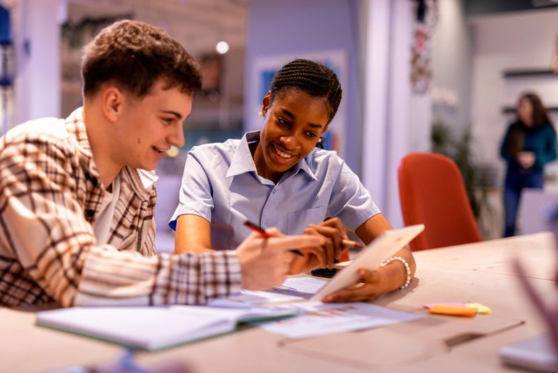 Two young students are smiling and working together on a project, using a digital tablet and taking notes, in a bright and modern classroom
